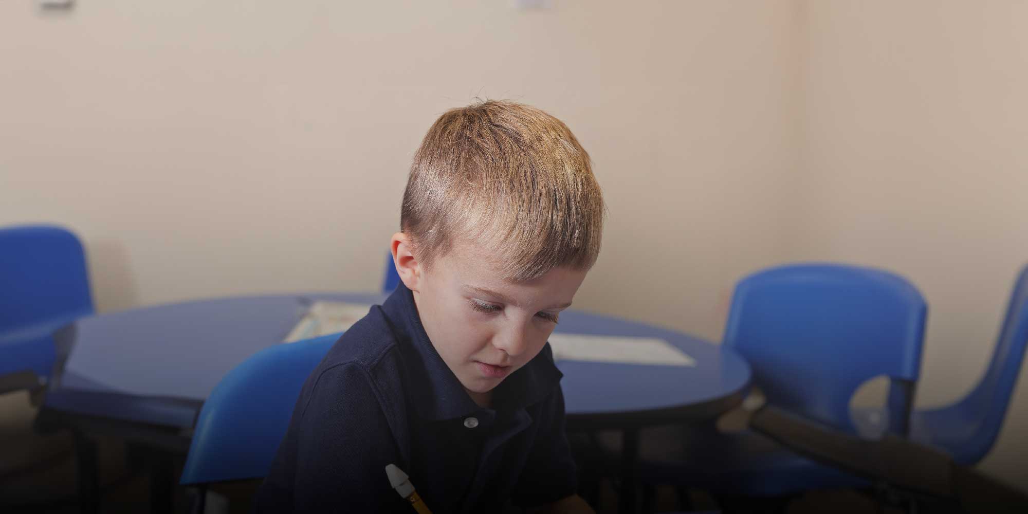 Young student writing with a pencil at a blue classroom table.